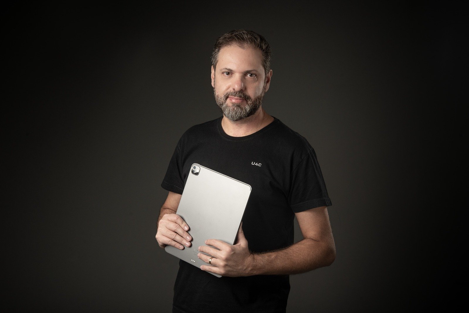 Túlio Iannini de camiseta preta da U4C, segurando um tablet, diante de um fundo escuro